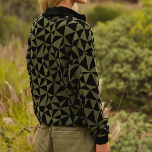 Kobieta o blond włosach, w pomarańczowych okularach przeciwsłonecznych, czarno-oliwkowym swetrze w geometryczne wzory i oliwkowych spodniach cargo stoi na zewnątrz w trawiastym, zielonym krajobrazie.