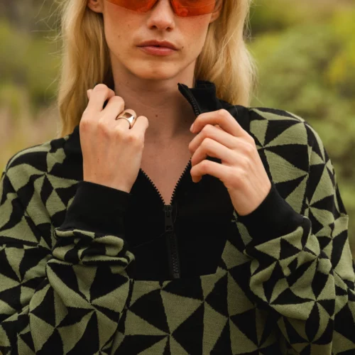 Kobieta z długimi blond włosami nosi pomarańczowe okulary przeciwsłoneczne i czarno-oliwkowy geometryczny wzór zapinany na zamek błyskawiczny, stojąc na zewnątrz z zielenią rozmytą w tle.