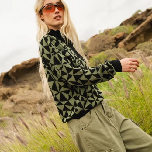 Kobieta z długimi blond włosami nosi pomarańczowe okulary przeciwsłoneczne, czarno-zielony sweter w geometryczne wzory i oliwkowe spodnie cargo, stojąc na zewnątrz w wysokiej trawie ze skalistym terenem w tle.