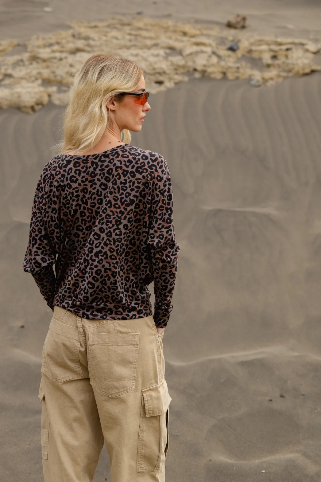 Osoba z długimi blond włosami w pomarańczowych okularach przeciwsłonecznych, brązowym topie w panterkę i beżowych spodniach cargo stoi na piaszczystym krajobrazie ze skałami w tle, odwrócona od kamery.