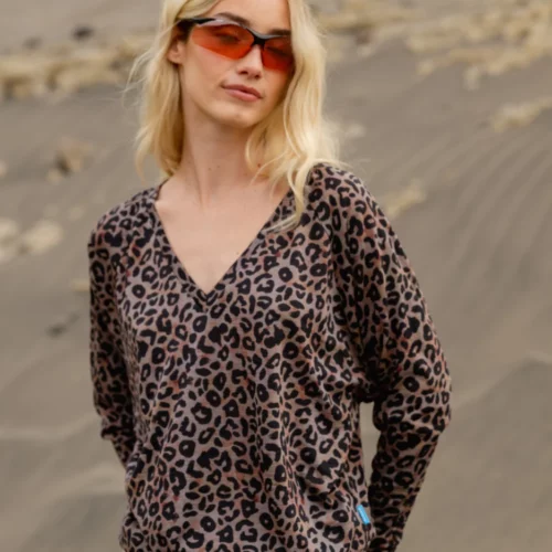 Kobieta o długich blond włosach, ubrana w czerwone okulary przeciwsłoneczne, bluzkę z dekoltem w serek w panterkę i jasnobeżowe spodnie stoi na zewnątrz w piaszczystym, skalistym krajobrazie.