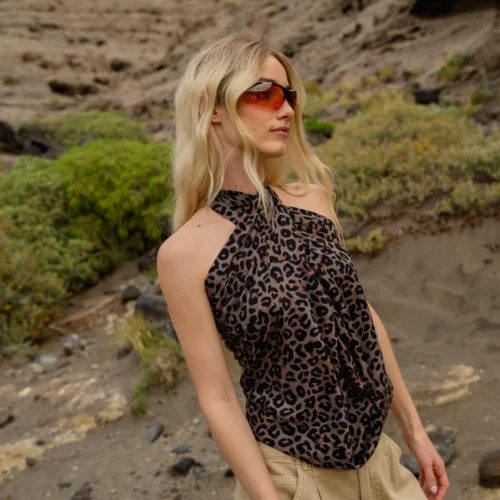 Kobieta o długich blond włosach w czerwonych okularach przeciwsłonecznych, bluzce bez rękawów w panterkę i brązowych spodniach cargo stoi na zewnątrz w skalistym, pustynnym krajobrazie z zielonymi krzewami.