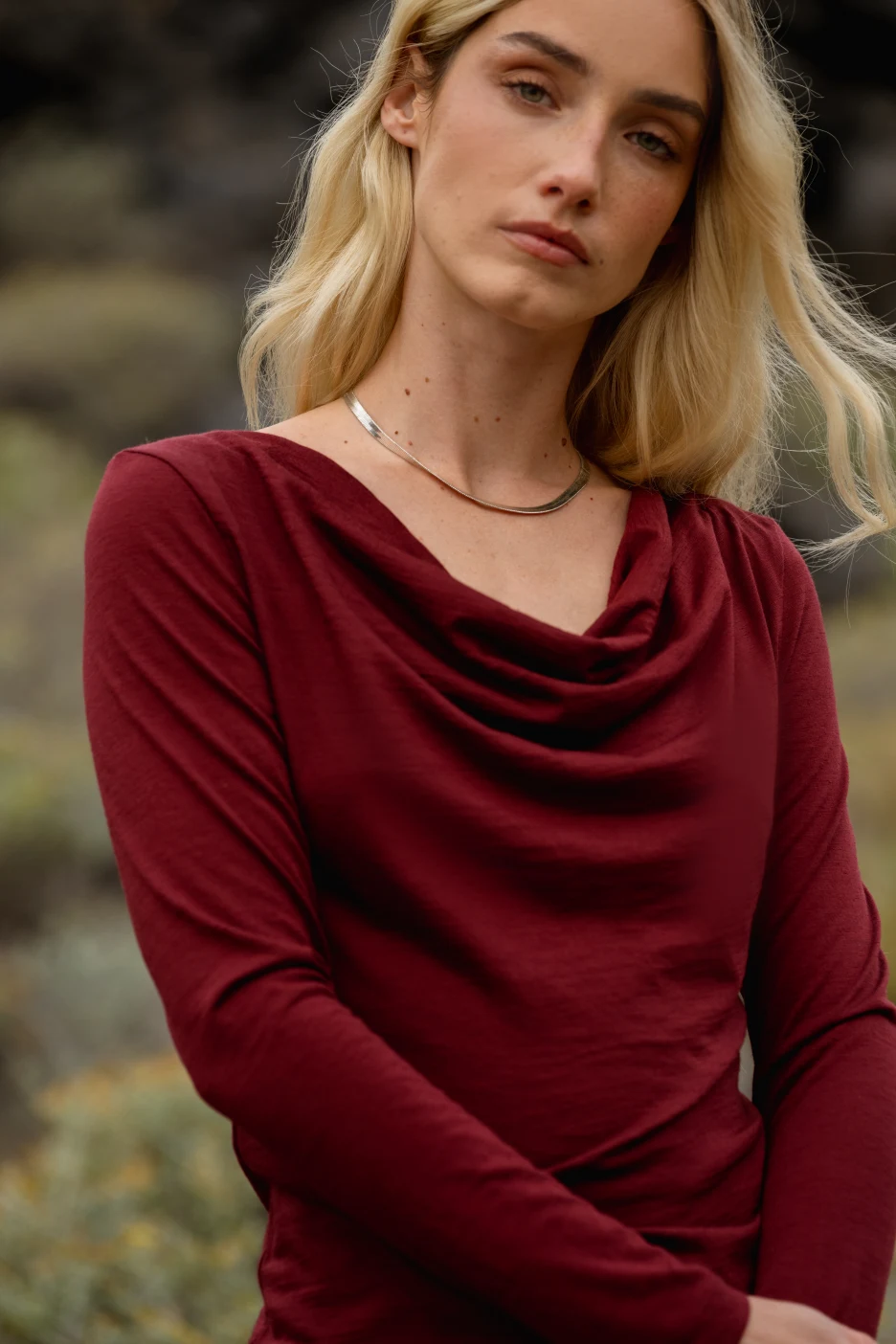 Kobieta z długimi blond włosami ubrana w bordową bluzkę z długim rękawem stoi na zewnątrz, patrząc lekko w bok z neutralnym wyrazem twarzy. Tło jest rozmyte naturalną zielenią.