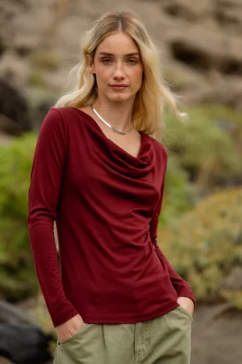 Kobieta z długimi blond włosami stoi na zewnątrz ubrana w bordową bluzkę z długimi rękawami i jasne spodnie, z rękami w kieszeniach. Tło jest rozmyte zielenią i skałami.
