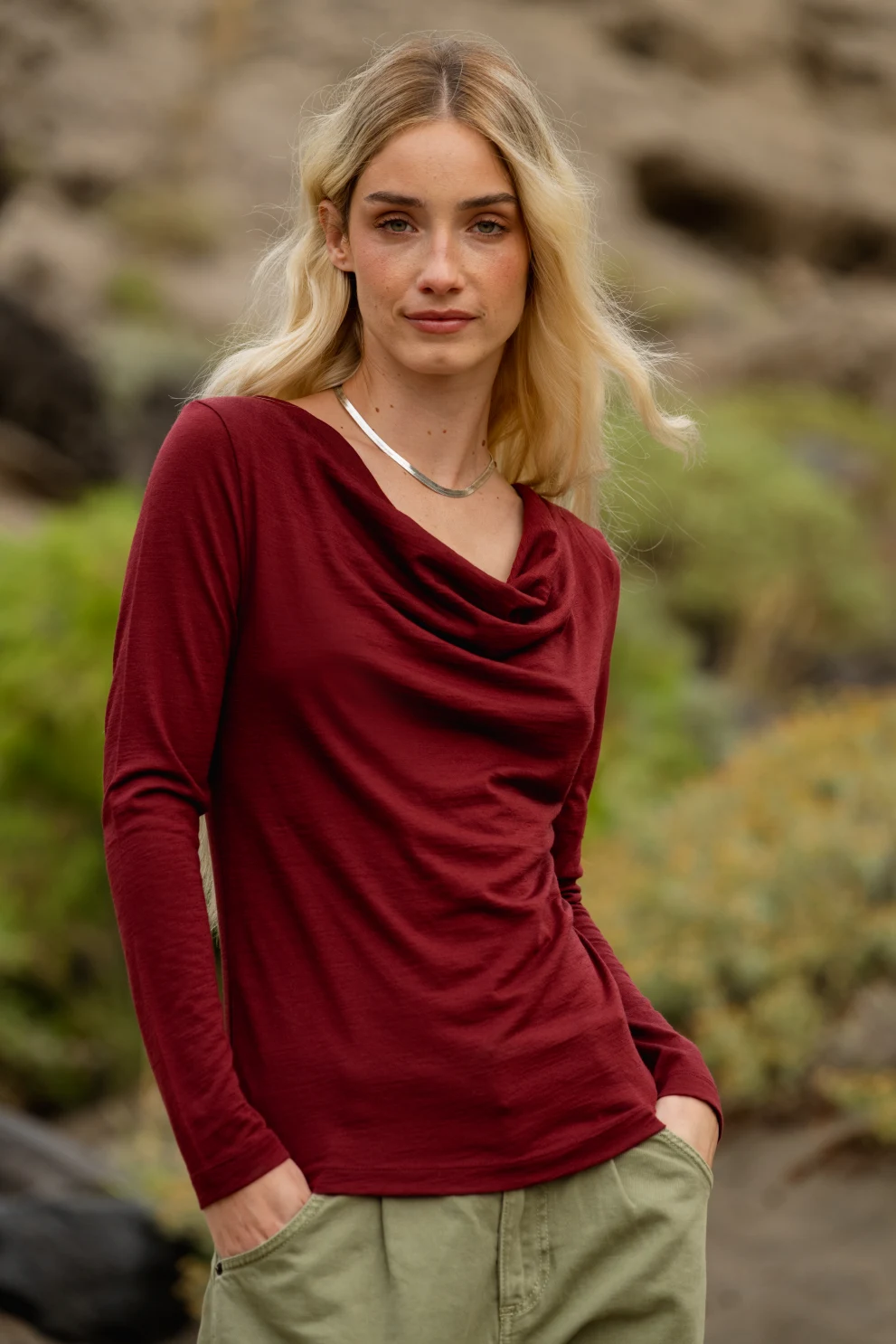 Kobieta z długimi blond włosami stoi na zewnątrz ubrana w bordową bluzkę z długimi rękawami i jasne spodnie, z rękami w kieszeniach. Tło jest rozmyte zielenią i skałami.