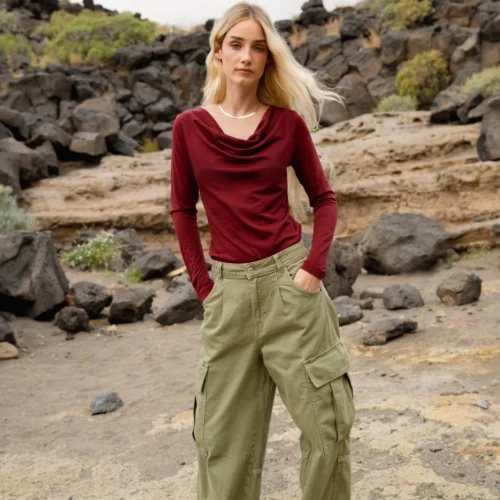 Kobieta z długimi blond włosami stoi na zewnątrz na skalistym terenie, ubrana w bordową bluzkę z długim rękawem, oliwkowozielone spodnie cargo i białe trampki. Stoi twarzą do kamery z rękami w kieszeniach, a w tle widać skały i krzewy.