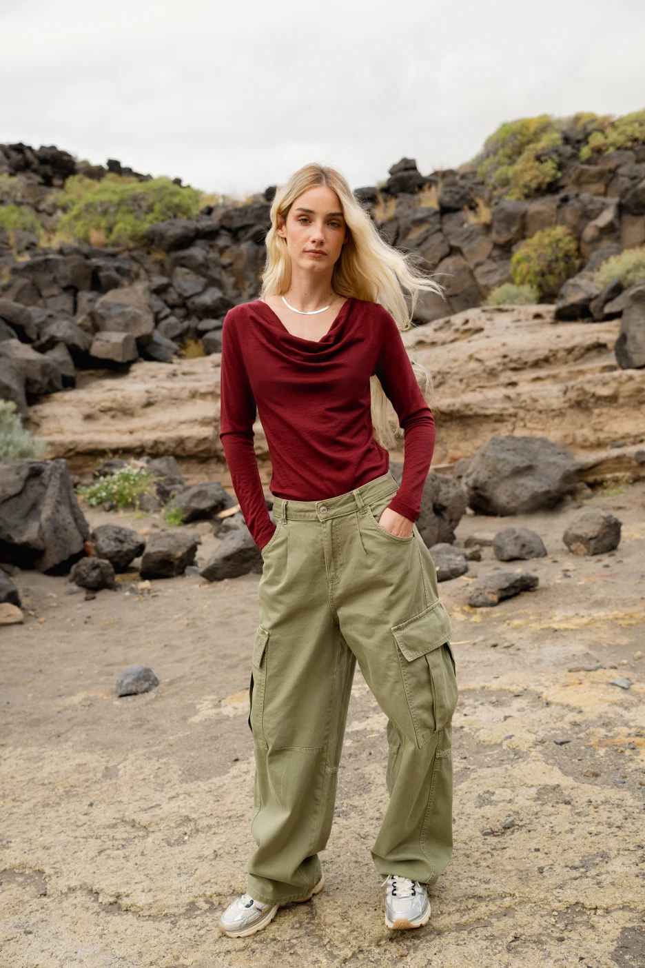 Kobieta z długimi blond włosami stoi na zewnątrz na skalistym terenie, ubrana w bordową bluzkę z długim rękawem, oliwkowozielone spodnie cargo i białe trampki. Stoi twarzą do kamery z rękami w kieszeniach, a w tle widać skały i krzewy.