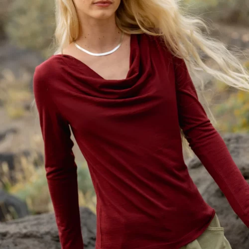 Kobieta z długimi blond włosami ma na sobie bordową bluzkę z długim rękawem i oliwkowozielone spodnie, stojąc na zewnątrz w pobliżu skał i liści.