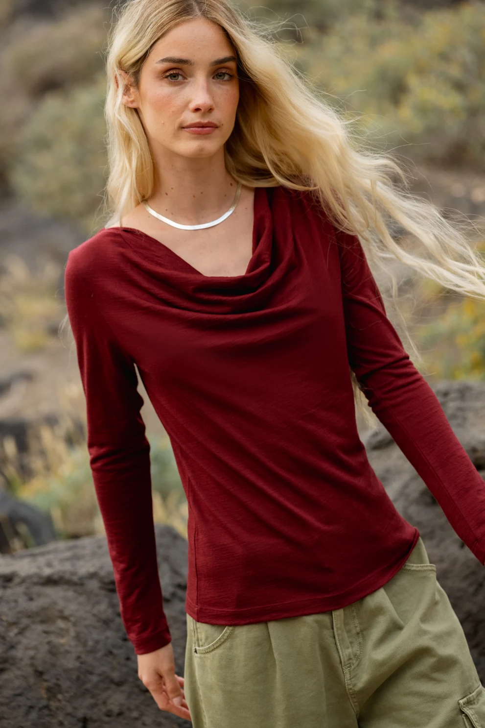 Kobieta z długimi blond włosami ma na sobie bordową bluzkę z długim rękawem i oliwkowozielone spodnie, stojąc na zewnątrz w pobliżu skał i liści.