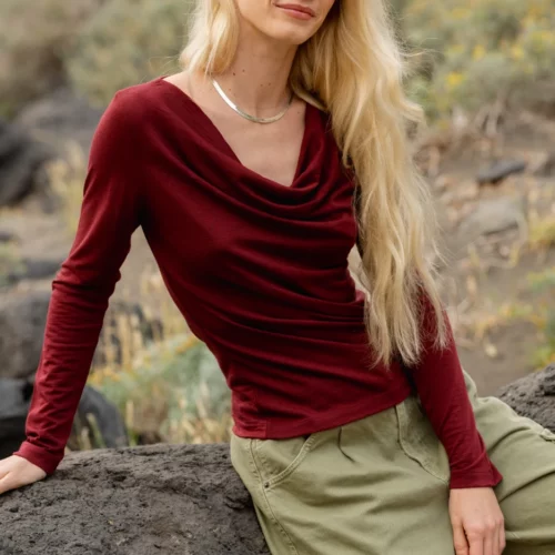 Kobieta z długimi blond włosami, ubrana w bordową bluzkę z długim rękawem i oliwkowozielone spodnie cargo, siedzi na dużej skale na zewnątrz, z suchą trawą i skałami w tle.