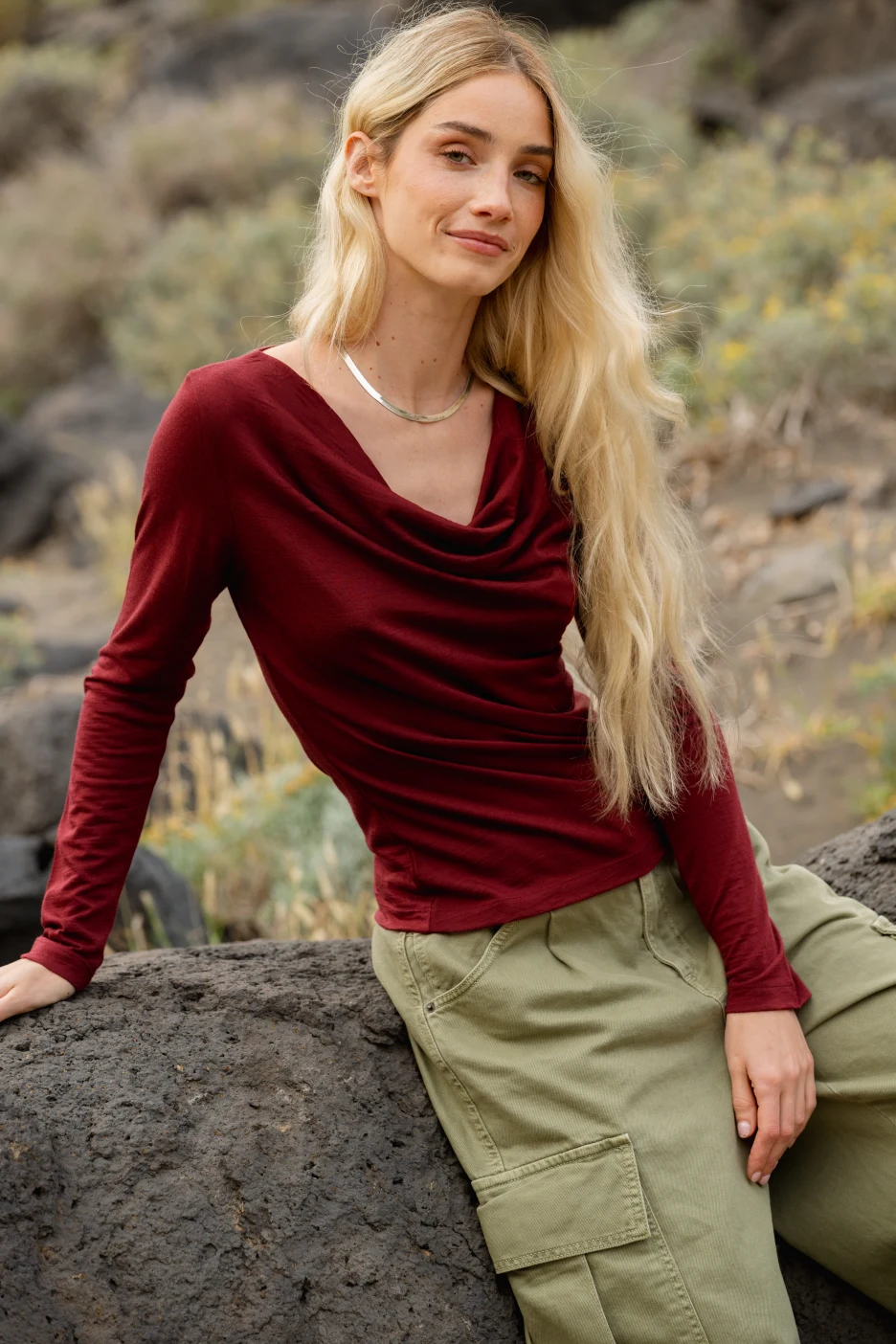 Kobieta z długimi blond włosami, ubrana w bordową bluzkę z długim rękawem i oliwkowozielone spodnie cargo, siedzi na dużej skale na zewnątrz, z suchą trawą i skałami w tle.