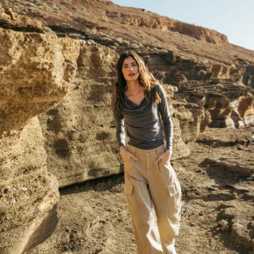 Kobieta ubrana w szarą bluzkę z długim rękawem i beżowe spodnie cargo spaceruje po skalistym, oświetlonym słońcem krajobrazie z brązowymi klifami w tle.
