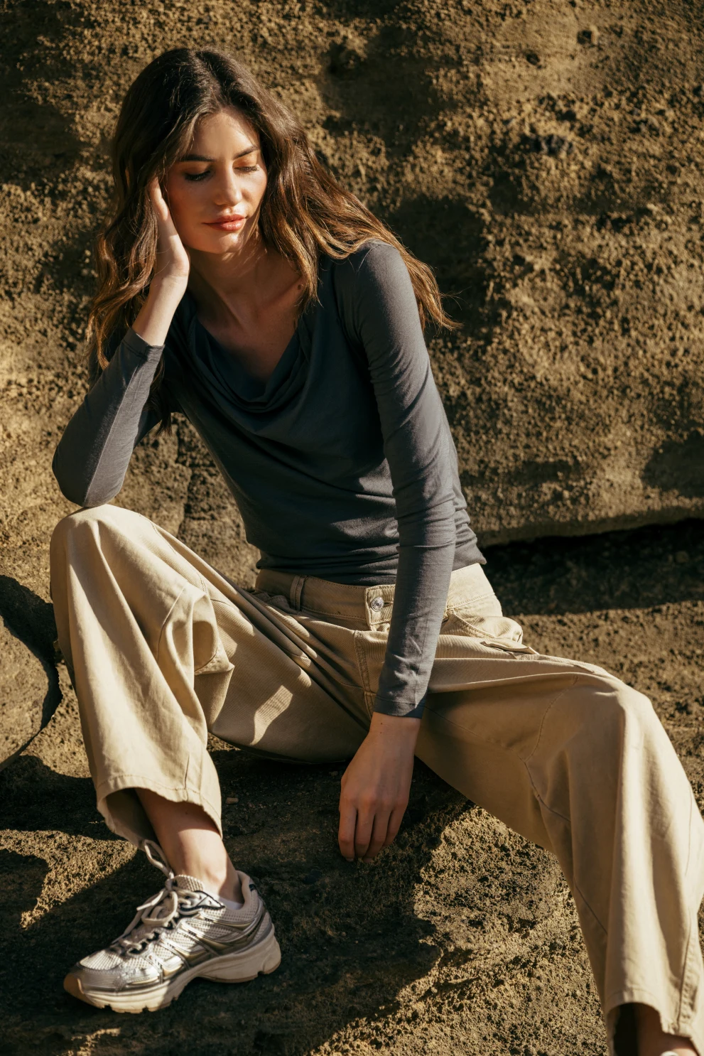 Kobieta z długimi brązowymi włosami siedzi na skale na zewnątrz, ubrana w szarą bluzkę z długim rękawem, beżowe spodnie z szerokimi nogawkami i białe trampki, a światło słoneczne rzuca cienie na skaliste tło.