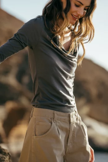 Kobieta z falującymi włosami ma na sobie szarą bluzkę z długim rękawem i beżowe spodnie, stojąc na zewnątrz z rozmytym skalistym terenem w tle. Uśmiecha się i patrzy w dół.