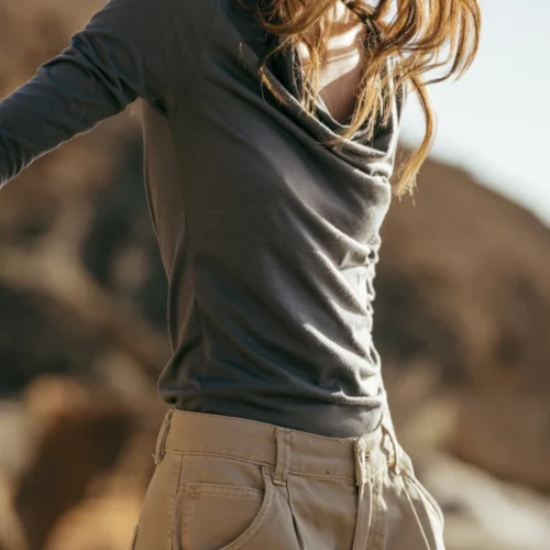 Kobieta z falującymi włosami ma na sobie szarą bluzkę z długim rękawem i beżowe spodnie, stojąc na zewnątrz z rozmytym skalistym terenem w tle. Uśmiecha się i patrzy w dół.