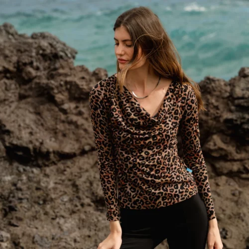 Kobieta w bluzce z długim rękawem w lamparcie cętki i czarnych spodniach stoi na ciemnym skalistym terenie nad oceanem, z falami i błękitną wodą w tle. Jest zwrócona lekko w bok z zamkniętymi oczami.