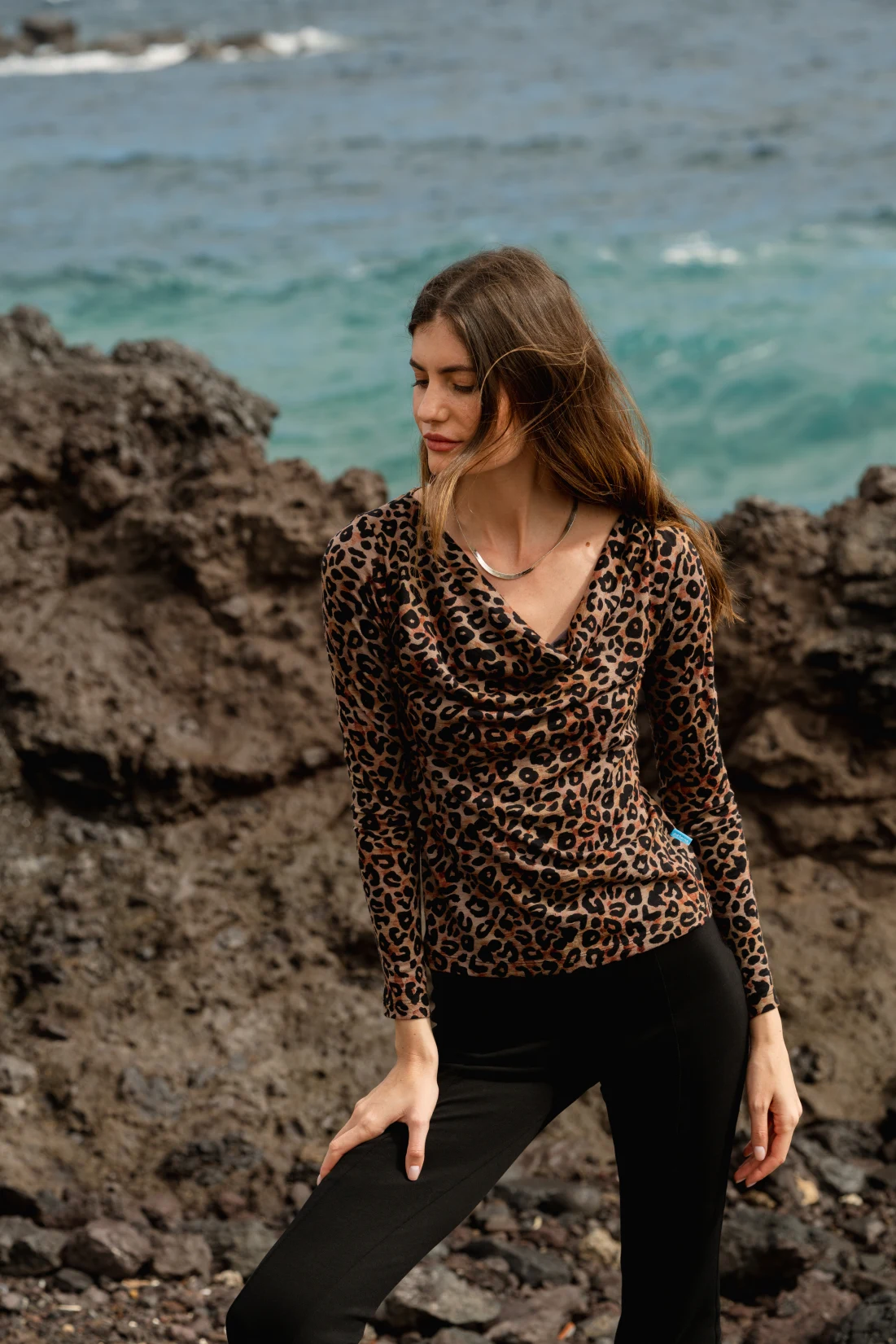 Kobieta w bluzce z długim rękawem w lamparcie cętki i czarnych spodniach stoi na ciemnym skalistym terenie nad oceanem, z falami i błękitną wodą w tle. Jest zwrócona lekko w bok z zamkniętymi oczami.