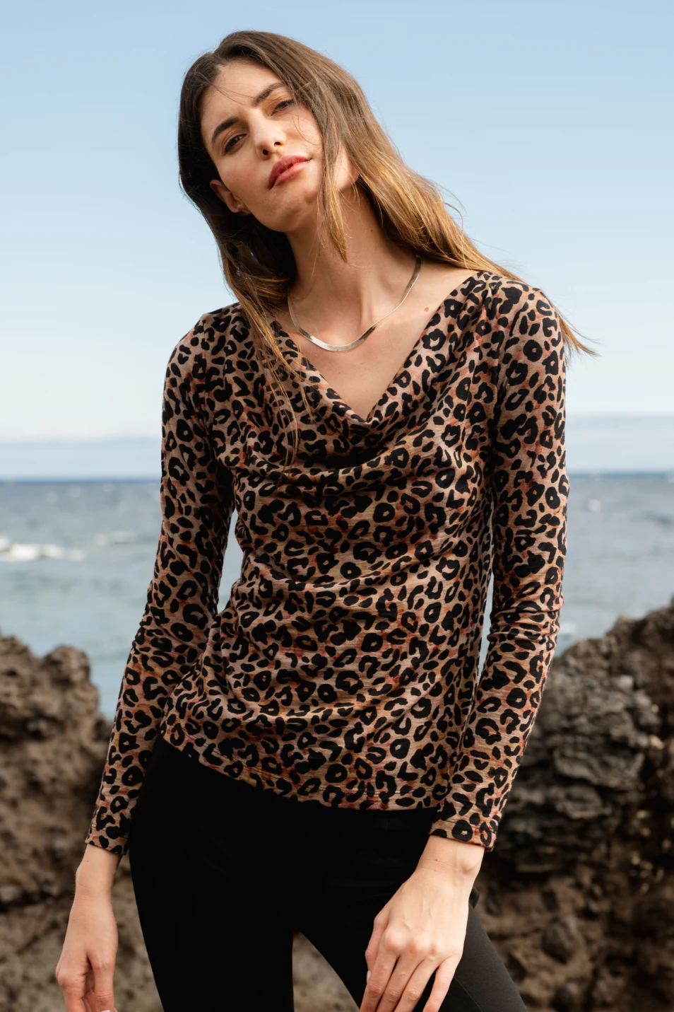Kobieta z długimi brązowymi włosami ma na sobie bluzkę z długim rękawem w lamparcie cętki i czarne spodnie, stojąc na zewnątrz w pobliżu skalistego terenu z oceanem i niebem w tle.