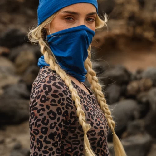 Kobieta z długimi blond warkoczami nosi niebieską chustę i osłonę twarzy oraz bluzkę w lamparcie cętki. Stoi na zewnątrz z rozmytymi skałami w tle, patrząc bezpośrednio w kamerę.