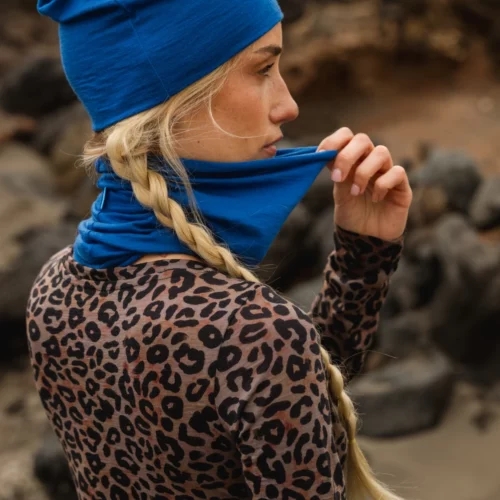 Osoba z długimi blond warkoczami w niebieskim kapeluszu i pasującym ochraniaczu na szyję, podnosi ochraniacz na twarz. Ubrana jest w bluzkę z nadrukiem lamparta i stoi na zewnątrz na skalistym terenie.