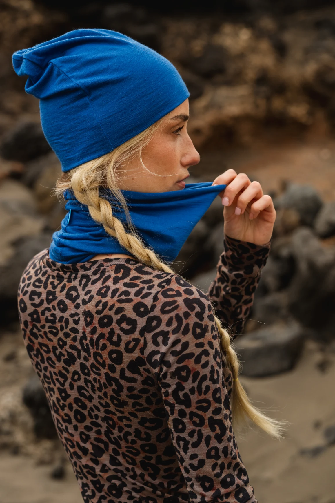 Osoba z długimi blond warkoczami w niebieskim kapeluszu i pasującym ochraniaczu na szyję, podnosi ochraniacz na twarz. Ubrana jest w bluzkę z nadrukiem lamparta i stoi na zewnątrz na skalistym terenie.