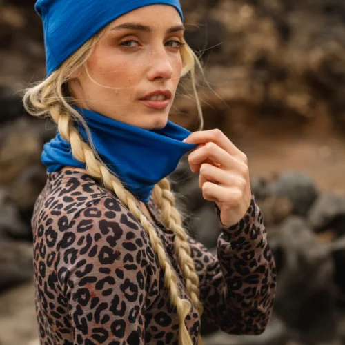 Kobieta z długimi blond warkoczami ma na sobie niebieską czapkę i ocieplacz na szyję, a także bluzkę z nadrukiem lamparta, stojąc na zewnątrz ze skałami rozmytymi w tle. Patrzy w stronę kamery, trzymając jedną ręką ocieplacz na szyję.