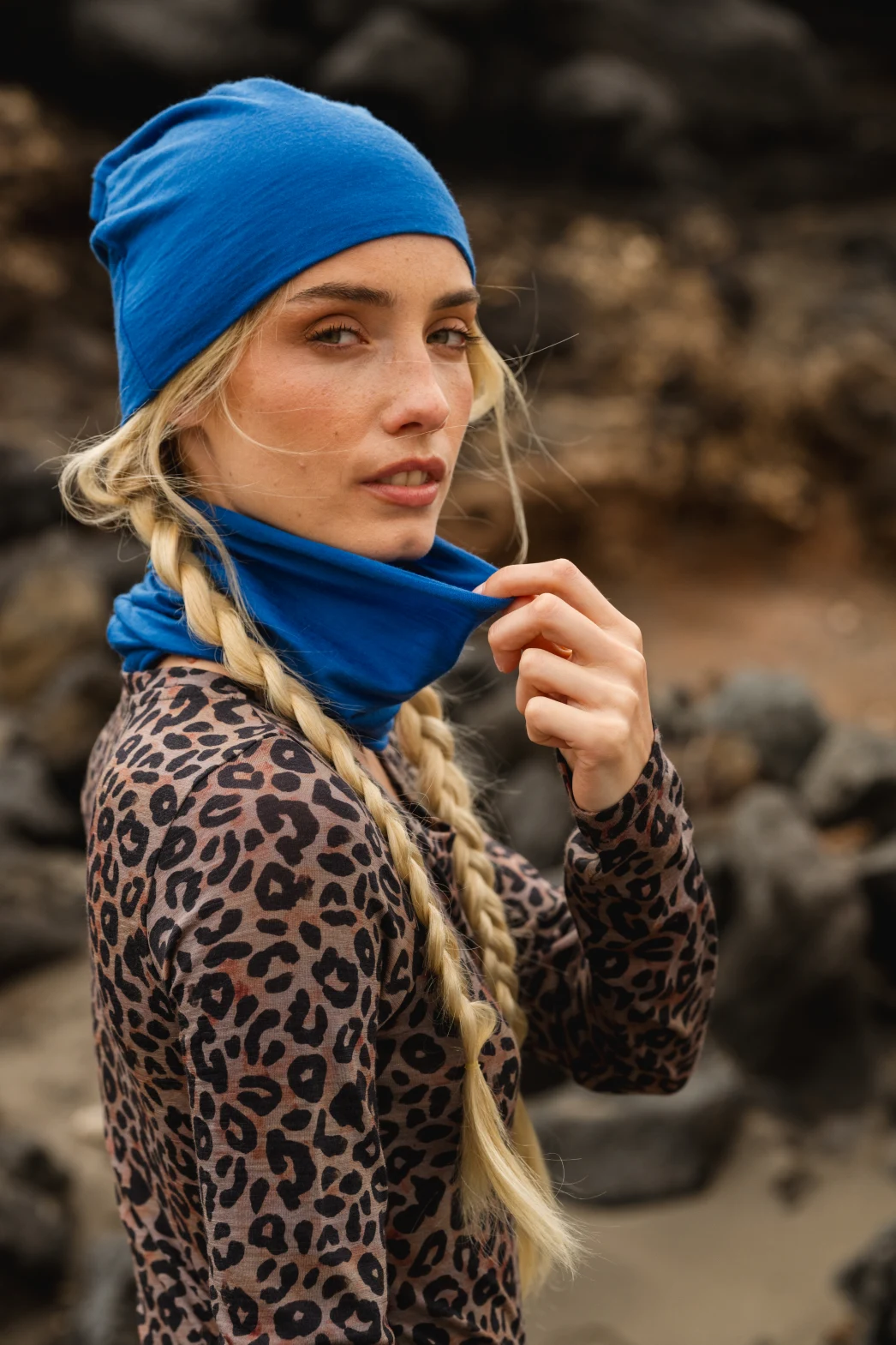 Kobieta z długimi blond warkoczami ma na sobie niebieską czapkę i ocieplacz na szyję, a także bluzkę z nadrukiem lamparta, stojąc na zewnątrz ze skałami rozmytymi w tle. Patrzy w stronę kamery, trzymając jedną ręką ocieplacz na szyję.