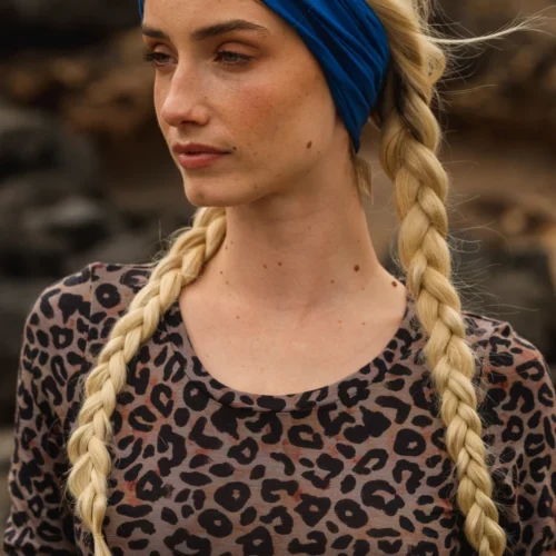 Kobieta z długimi blond warkoczami i niebieską opaską na głowie, ubrana w bluzkę w panterkę, stoi na zewnątrz z neutralnym wyrazem twarzy i skalistym tłem.