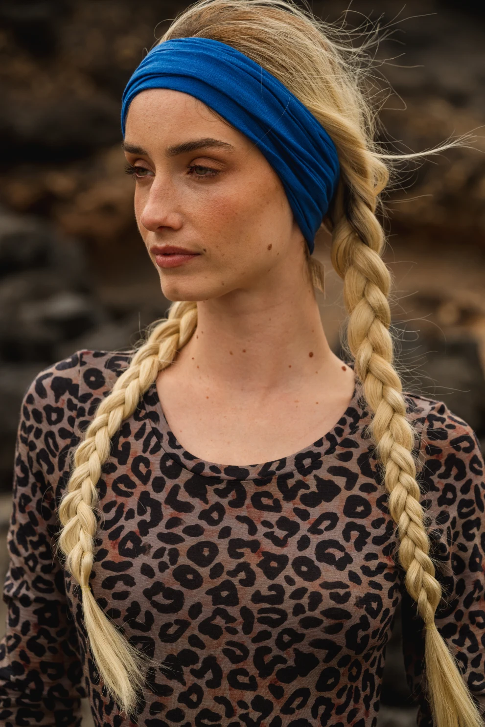 Kobieta z długimi blond warkoczami i niebieską opaską na głowie, ubrana w bluzkę w panterkę, stoi na zewnątrz z neutralnym wyrazem twarzy i skalistym tłem.
