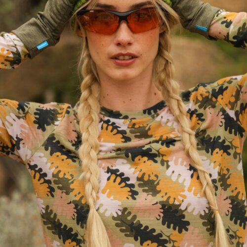 Kobieta z długimi blond warkoczami nosi zieloną czapkę, pomarańczowe okulary przeciwsłoneczne, oliwkowe rękawiczki i koszulę w kamuflażowe wzory, stojąc na zewnątrz z rękami na kapeluszu.