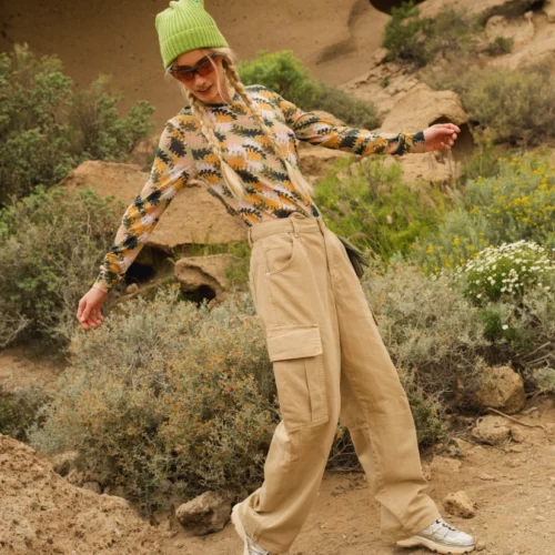 Osoba na zewnątrz ubrana w beżowe spodnie cargo, kolorową koszulę z długim rękawem, zieloną czapkę, okulary przeciwsłoneczne i trampki, stojąca na kamienistej ścieżce z krzewami i dużymi formacjami skalnymi w tle.
