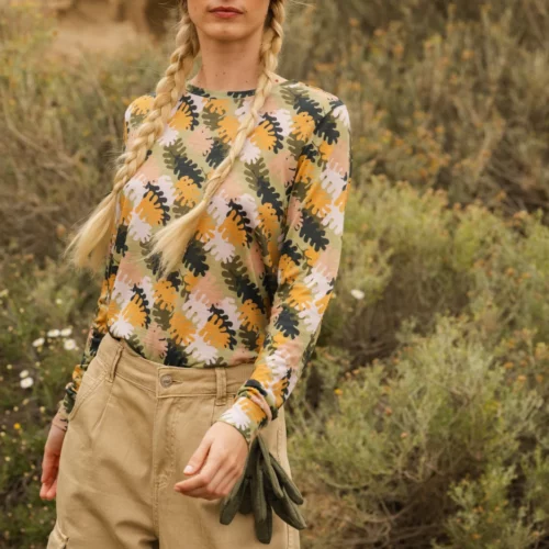 Osoba z blond warkoczami ma na sobie zieloną czapkę, duże pomarańczowe okulary przeciwsłoneczne, wzorzystą koszulę z długim rękawem i brązowe spodnie cargo, stojąc na zewnątrz w naturalnym, skalistym otoczeniu z krzewami.