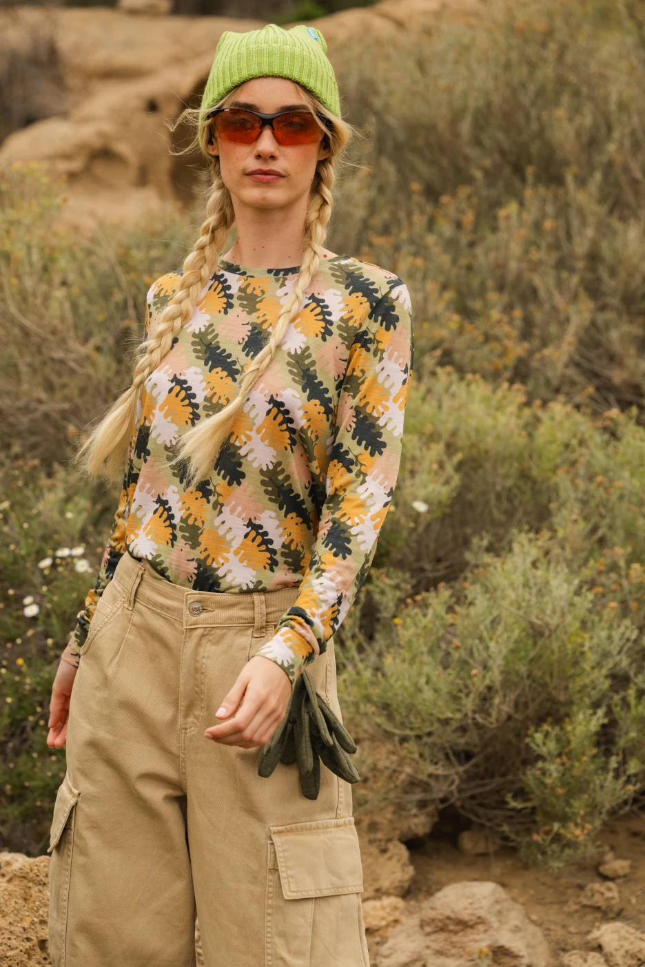 Osoba z blond warkoczami ma na sobie zieloną czapkę, duże pomarańczowe okulary przeciwsłoneczne, wzorzystą koszulę z długim rękawem i brązowe spodnie cargo, stojąc na zewnątrz w naturalnym, skalistym otoczeniu z krzewami.