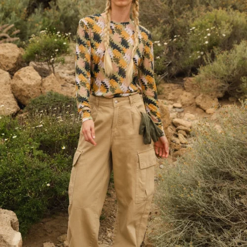 Osoba stoi na zewnątrz w trudnym terenie, ubrana w zieloną dzianinową czapkę, pomarańczowe okulary przeciwsłoneczne, wzorzystą koszulę z długim rękawem, workowate spodnie cargo w kolorze khaki i srebrne trampki. Włosy są splecione w dwa długie warkocze.