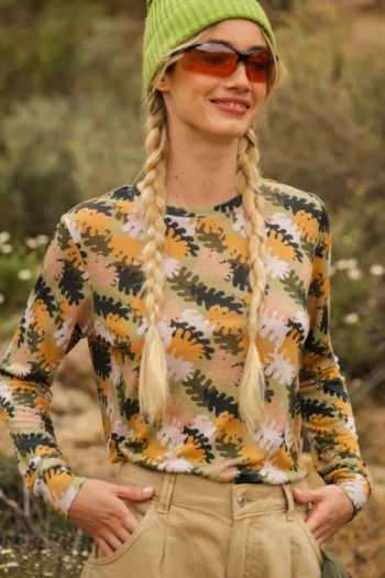 Kobieta z długimi blond warkoczami ma na sobie zieloną czapkę, pomarańczowe okulary przeciwsłoneczne, wielokolorową kamuflażową koszulę z długim rękawem i brązowe spodnie, stojąc na zewnątrz z zielenią w tle.