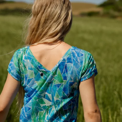Kobieta o długich blond włosach stoi na trawiastym polu, odwrócona tyłem, ubrana w koszulkę z wełny merino damskiej w kolorze niebieskim z dekoltem w kształcie litery V z tyłu. Czyste niebo i lekko rozmyte tło dopełniają scenę.