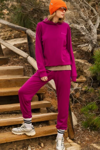 Osoba ubrana w jasny, purpurowy dres, pomarańczową czapkę, okulary przeciwsłoneczne i białe trampki stoi na drewnianych schodach na zewnątrz, otoczona drzewami i liśćmi.