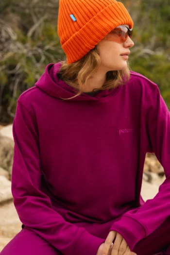 Osoba ubrana w pomarańczową czapkę, okulary przeciwsłoneczne i magentową bluzę z kapturem siedzi na zewnątrz, patrząc w bok. Zieleń i skały są widoczne w rozmytym tle.