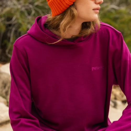 Osoba ubrana w pomarańczową czapkę, okulary przeciwsłoneczne i magentową bluzę z kapturem siedzi na zewnątrz, patrząc w bok. Zieleń i skały są widoczne w rozmytym tle.