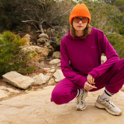 Osoba w karmazynowym dresie, pomarańczowej czapce i okularach przeciwsłonecznych przysiada na skalistej ścieżce otoczonej zielenią i drzewami, w zrelaksowanej pozie i neutralnym wyrazie twarzy.