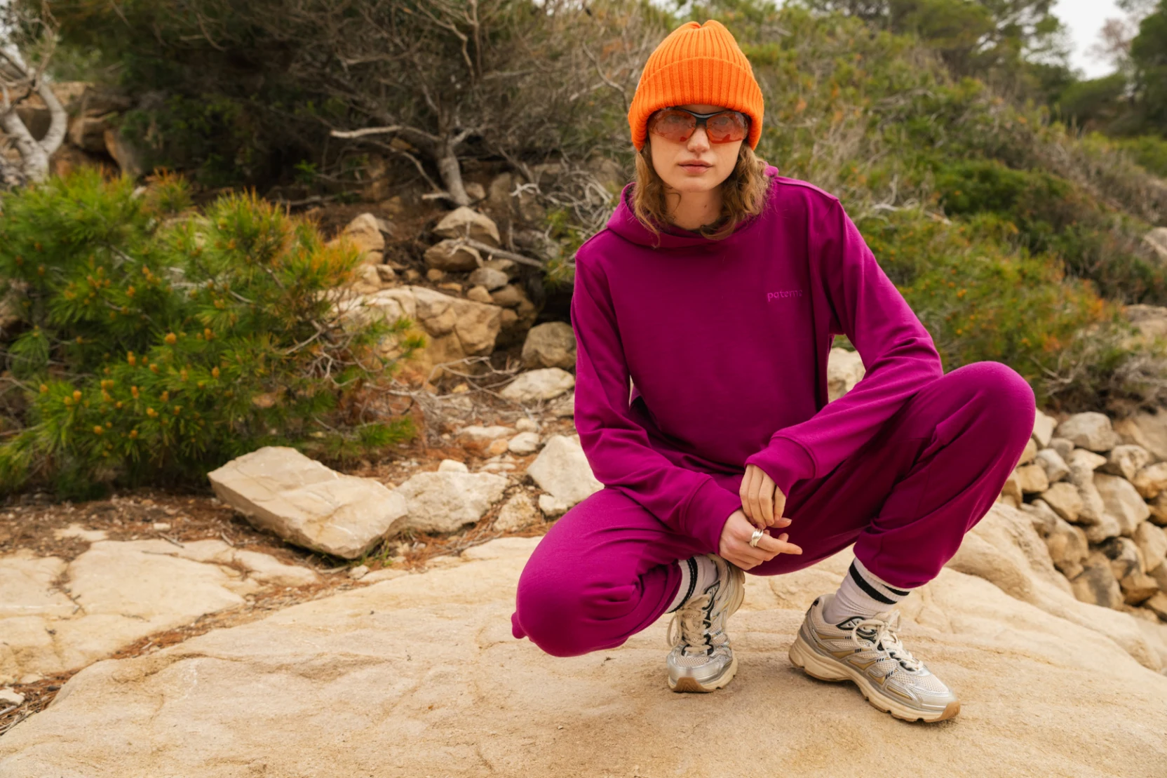 Osoba w karmazynowym dresie, pomarańczowej czapce i okularach przeciwsłonecznych przysiada na skalistej ścieżce otoczonej zielenią i drzewami, w zrelaksowanej pozie i neutralnym wyrazie twarzy.