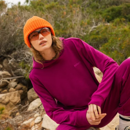 Osoba ubrana w pomarańczową czapkę, pomarańczowe okulary przeciwsłoneczne i karmazynowy dres przykuca na zewnątrz na skalistym terenie z zielenią w tle.