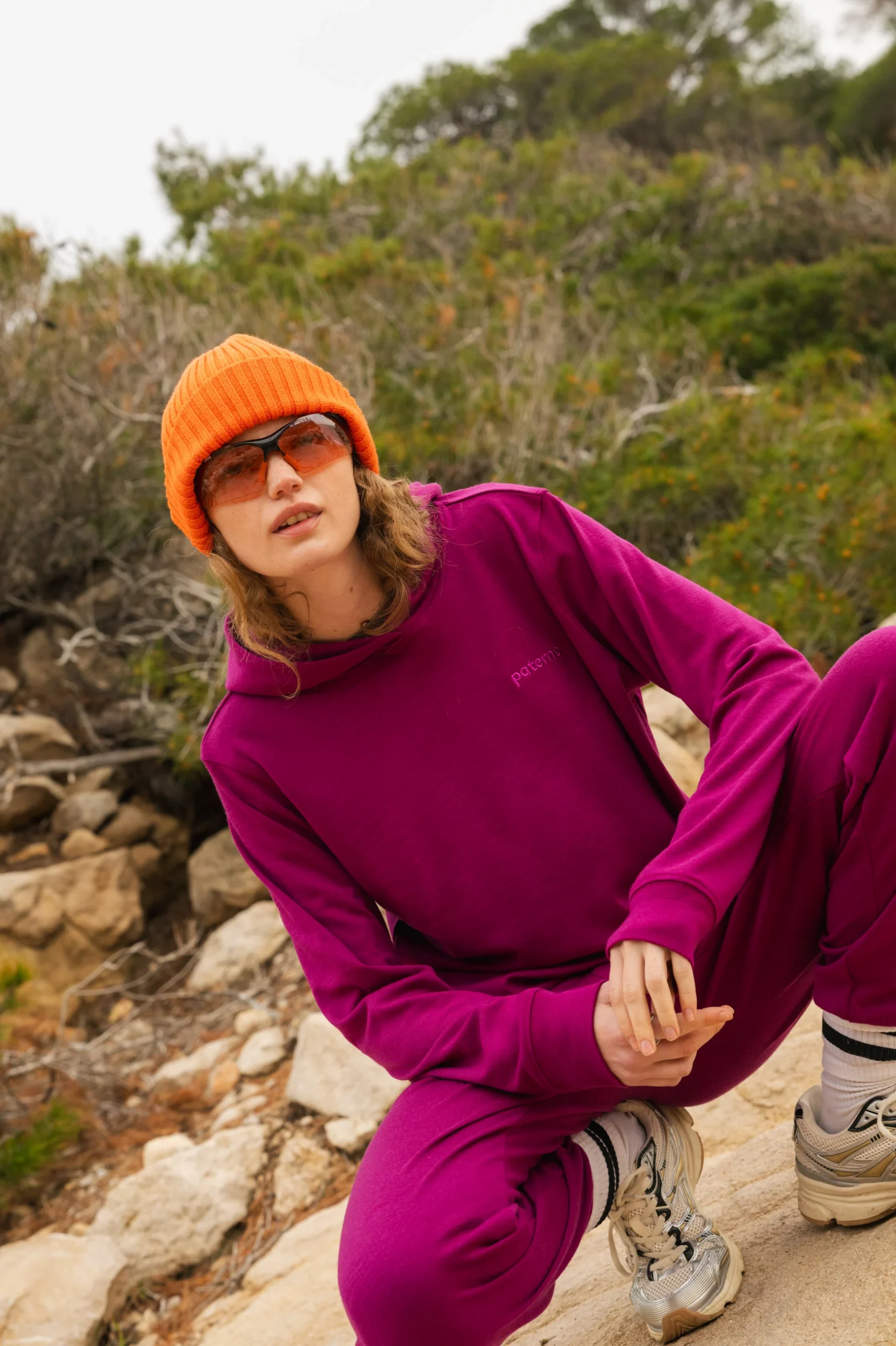 Osoba ubrana w pomarańczową czapkę, pomarańczowe okulary przeciwsłoneczne i karmazynowy dres przykuca na zewnątrz na skalistym terenie z zielenią w tle.