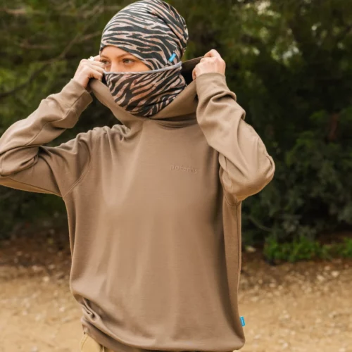 Osoba na zewnątrz podnosi szyję brązowego swetra na część twarzy, nosząc wzorzystą czapkę w paski przypominające zebrę. W tle widoczne są zielone drzewa i ziemia.