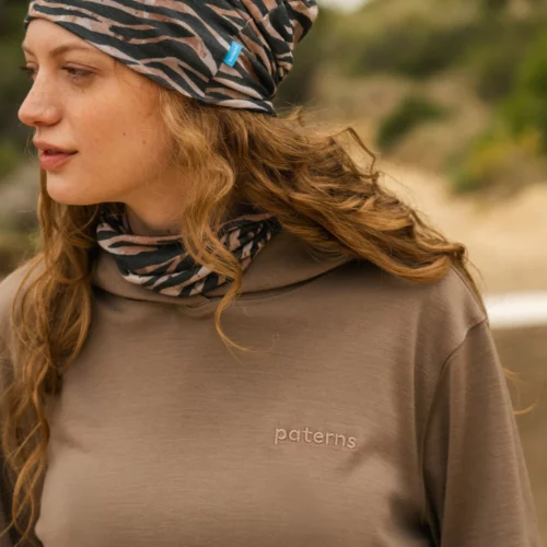 Kobieta z falującymi włosami ma na sobie brązową wzorzystą chustę i pasujący ochraniacz na szyję, a także brązową koszulę z wyhaftowanymi paternami. Stoi na zewnątrz z drzewami i piaskiem w tle.