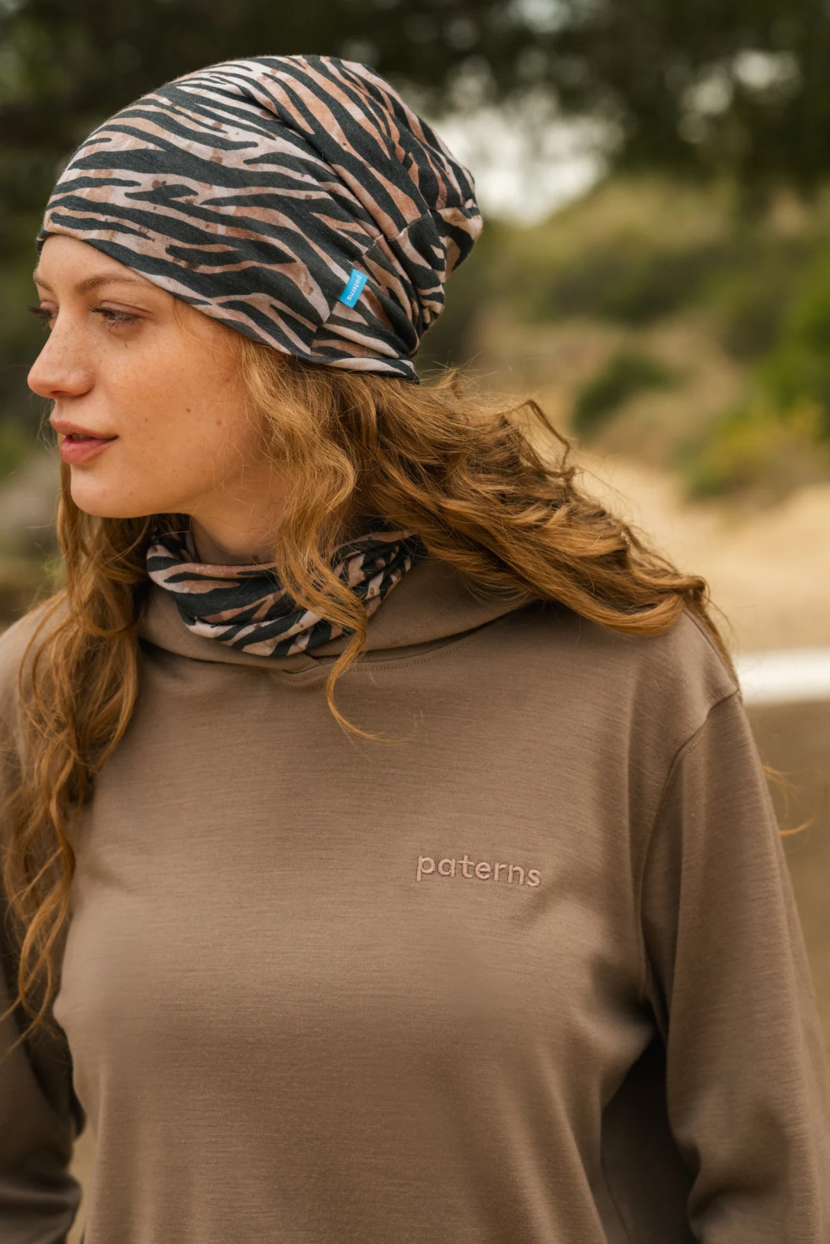 Kobieta z falującymi włosami ma na sobie brązową wzorzystą chustę i pasujący ochraniacz na szyję, a także brązową koszulę z wyhaftowanymi paternami. Stoi na zewnątrz z drzewami i piaskiem w tle.