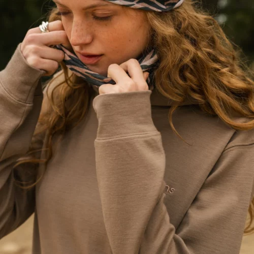 Młoda kobieta z długimi, falującymi włosami dopasowuje wzorzystą opaskę na głowę i pasujący ochraniacz na szyję. Ma na sobie luźną, beżową bluzę i stoi na zewnątrz, patrząc w dół ze spokojnym wyrazem twarzy.