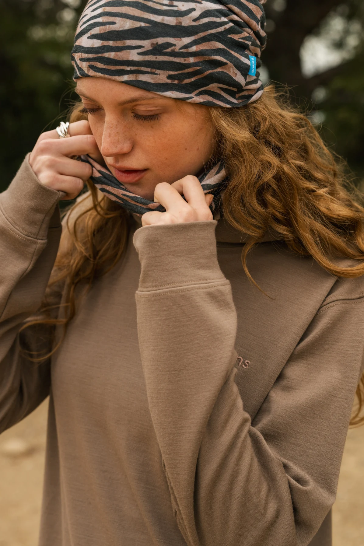 Młoda kobieta z długimi, falującymi włosami dopasowuje wzorzystą opaskę na głowę i pasujący ochraniacz na szyję. Ma na sobie luźną, beżową bluzę i stoi na zewnątrz, patrząc w dół ze spokojnym wyrazem twarzy.