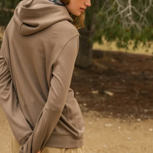 Osoba ubrana w ciemnoszarą bluzę z kapturem, brązowe spodnie cargo i chustę we wzór zebry stoi na zewnątrz na polnej ścieżce, patrząc w dół i w bok z rękami splecionymi za plecami. W tle widać drzewa.