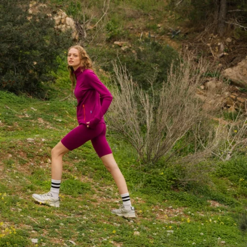 Kobieta ubrana w karmazynowy strój sportowy i białe trampki idzie pod górę trawiastą, kamienistą ścieżką otoczoną zielonymi krzewami i nielicznymi drzewami.